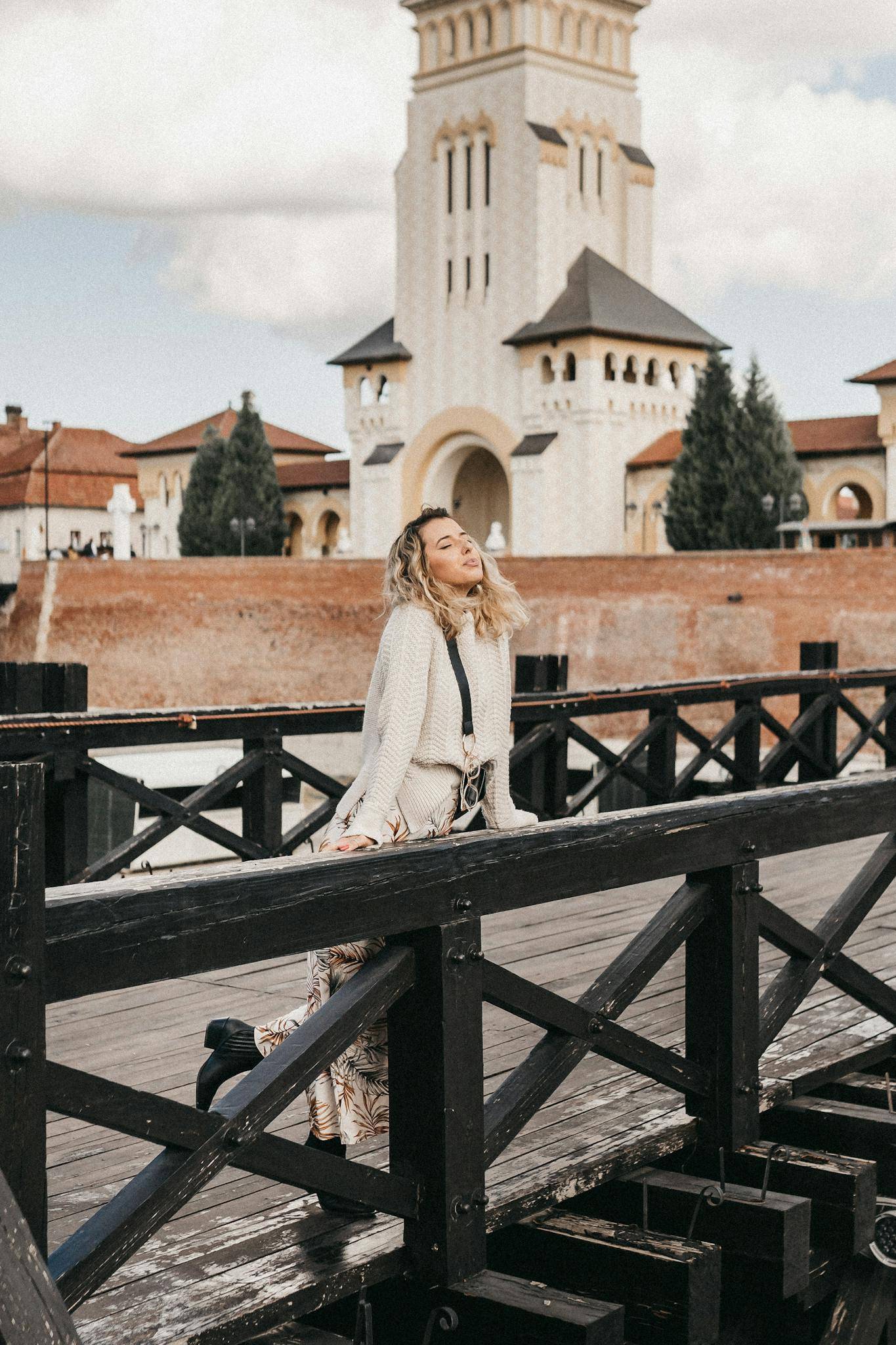 Full body of pleased female traveler leaning on railing of footbridge while standing on street with aged building in city